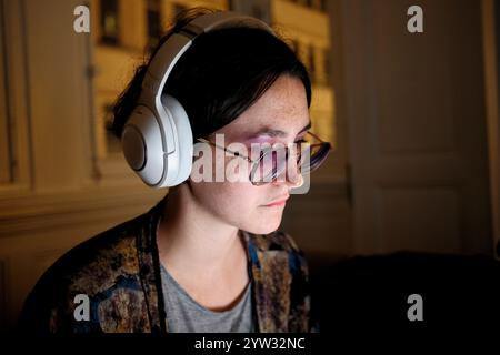 Eine fokussierte Frau mit Kopfhörern und Brillen schaut auf einen Computerbildschirm in einem schwach beleuchteten Raum. Stockfoto