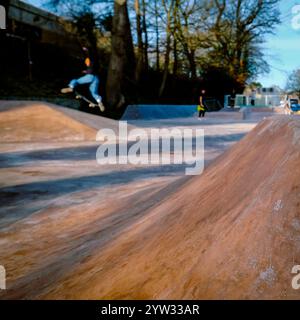 Ein Skateboarder mitten in der Luft macht einen Trick im Kingsbridge Skatepark an einem sonnigen Tag, umgeben von Bäumen, geparkten Autos und urbaner Landschaft Stockfoto