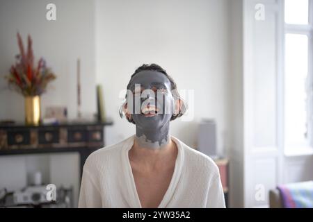 Eine junge Frau mit einer Gesichtsmaske, die in einem hellen, gemütlichen Zimmer lächelt. Stockfoto