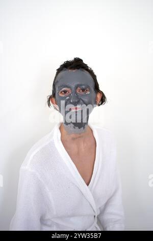 Eine junge Frau mit einer Gesichtsmaske, die in einem weißen Gewand vor einem schlichten Hintergrund posiert. Stockfoto