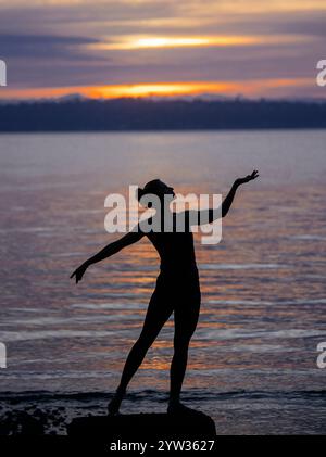 Silhouette einer Frau, die Ballett am Wasser bei Sonnenuntergang übt. Stockfoto