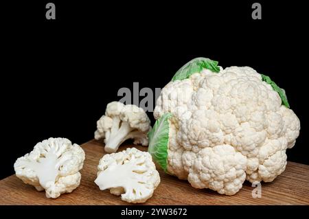 Ein frischer Blumenkohlkopf wird auf ein hölzernes Schneidebrett mit gehackten Röschen vor schwarzem Hintergrund gelegt. Stockfoto