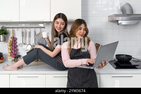 Zwei Frauen mit gestreifter Schürze lesen oder mit Laptop-Bildschirm in einer modernen Küche interagieren. Kochutensilien stehen auf der Arbeitsplatte. Stockfoto