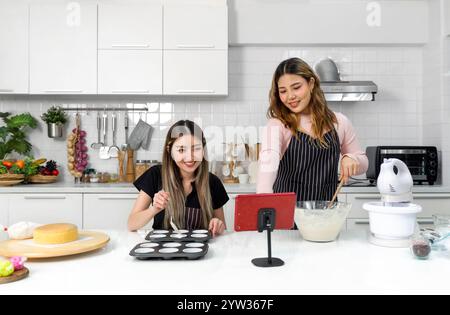 Zwei junge Frauen auf einer Schürze bereiten in der Küche Nachtisch zu, während sie auf dem Tablet-Computer im Online-Rezeptbuch nachschauen. Stockfoto