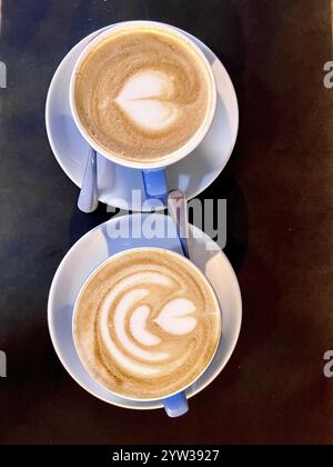 Blick von oben auf zwei Cappuccinos mit herzförmiger Latte Art, serviert in blauen Tassen auf einem dunklen Tisch. Stockfoto