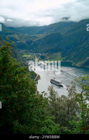 Majestätischer Fjord mit einem Kreuzfahrtschiff in der Nähe eines malerischen Dorfes, umgeben von üppigen grünen Bergen unter einem bewölkten Himmel, Geirangerfjord, Norwegen Stockfoto