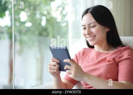 Glückliche asiatische Frau, die zu Hause auf einer Couch sitzt, hat ein eBook gepackt Stockfoto