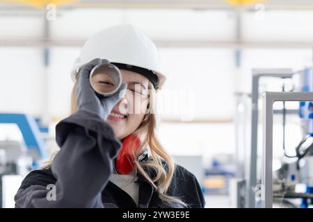 Lächelnde Ingenieurin mit Schutzhelm, die durch eine Metallstange in einer industriellen Umgebung schaut, Deutschland Stockfoto