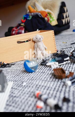 Ein Teddybär lehnt sich an eine hölzerne Spielzeugkiste, inmitten verstreuter Spielzeuge auf einem gemusterten Teppich, Deutschland Stockfoto