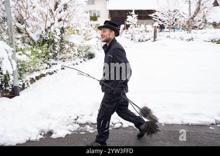 Ein Schornsteinfeger in dunkler Arbeitskleidung spaziert entlang einer schneebedeckten Straße. Er trägt seine Reinigungswerkzeuge. Die Szene deutet auf einen kalten Wintertag hin. Stockfoto