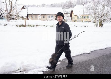 Ein fröhlicher Schornsteinfeger geht selbstbewusst durch eine schneebedeckte Dorfstraße und trägt seine Putzwerkzeuge. Die heitere Winterszene zeigt ein Q Stockfoto