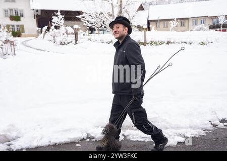 Ein fröhlicher Schornsteinfeger geht selbstbewusst durch eine schneebedeckte Dorfstraße und trägt seine Putzwerkzeuge. Die heitere Winterszene zeigt ein Q Stockfoto