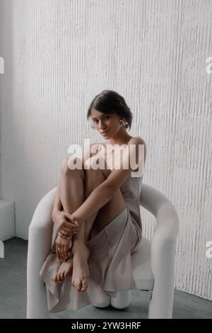 Die junge Frau sitzt anmutig in einem modernen Sessel und trägt ein zartes Kleid. Minimalistischer Hintergrund betont ihr ausgeglichenes Auftreten und ihre subtile Eleganz A Stockfoto