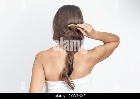 Braunes langes Naturhaar, zu Flecht verdreht. Kämmen mit Kamm. Frau im Handtuch auf weiß. Rückansicht Stockfoto