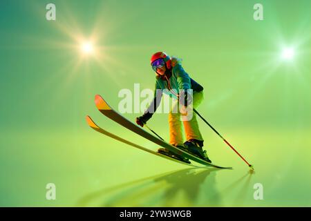 Weibliche Skiläuferin mit dynamischer Abfahrt im grünen Neonlicht vor leuchtendem grünem Hintergrund mit Sonnenstrahlen. Stockfoto