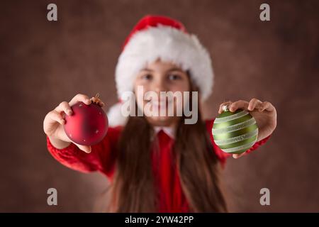 Lächelndes Mädchen hält Weihnachtsbaumkugeln Dekoration in seinen Augen und trägt Santa Claus Hut Kleid Porträt auf rotem Hintergrund Stockfoto