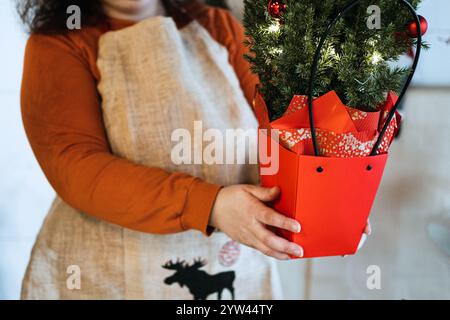 Hände, die einen kleinen Tannenbaum mit Ornamenten und Bändern dekorieren. Mit personalisiertem Weihnachtsgebäck und handgefertigten Geschenkideen. Stockfoto