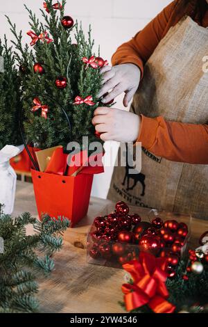 Hände, die einen kleinen Tannenbaum mit Ornamenten und Bändern dekorieren. Mit personalisiertem Weihnachtsgebäck und handgefertigten Geschenkideen. Stockfoto