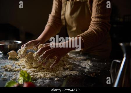 Eine Person, die eine Schürze trägt, knetscht Teig auf einer bemehlten Oberfläche, wodurch eine rustikale Atmosphäre entsteht. Frische Zutaten und natürliches Licht unterstreichen die Tradition Stockfoto