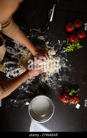 Blick von oben auf die Hände, die Mehl zu Teig kneten, umgeben von frischen Tomaten und Kräutern. Die Szene erinnert an hausgemachte Küche und kulinarische Kreativität, perfekt Stockfoto