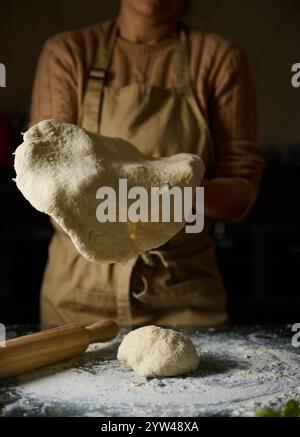 Eine Person, die eine Schürze trägt, dehnt selbstgemachten Teig auf einer bemehlten Oberfläche aus, bereit zum Backen. Die warme Beleuchtung vermittelt eine gemütliche, intime Küche Atmosphäre Stockfoto