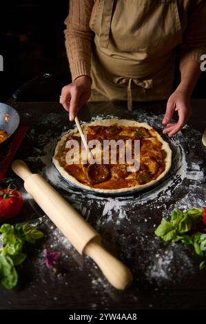 Eine Person in einer Schürze verteilt Pizzasauce auf der Kruste. Zutaten und Utensilien umgeben sie und verbessern das hausgemachte kulinarische Erlebnis. Stockfoto