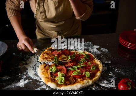 Nahaufnahme einer Person, die eine frisch gebackene, hausgemachte Pizza mit Tomaten und Basilikum auf einer bemehlten Küchenfläche schneidet. Stockfoto