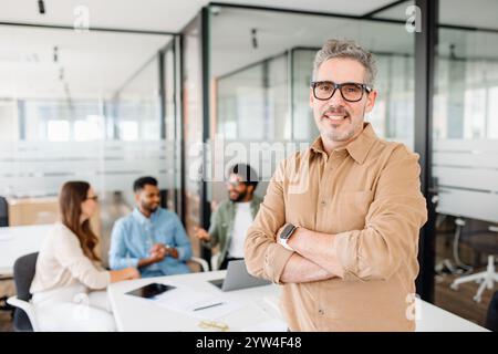 Ein reifer, hochqualifizierter Mann, Manager oder Start-up-Inhaber, steht stolz vor einem modernen Büroumfeld mit überkreuzten Armen, während sein Team hinter ihm eine lebhafte Diskussion führt, ein effektives Führungskonzept Stockfoto