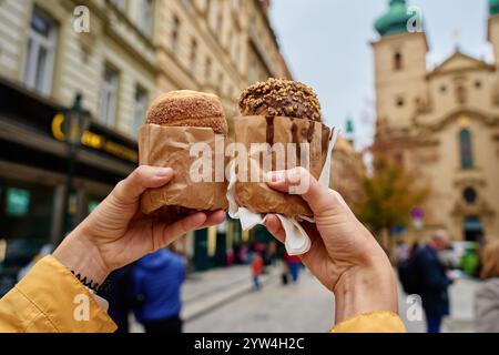 Nahaufnahme einer Frau, die zwei Kaminkuchen in Papier mit verschwommener Stadt im Hintergrund hält. Touristen genießen frisch gebackenen Trdelnik auf der Straße in Pra Stockfoto