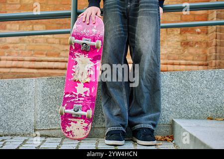 Skateboarder in Baggy-Jeans mit einem zerkratzten rosa Skateboard auf der City Street. Der Teenager ist Skateboarding. Konzept der Jugendsubkultur Stockfoto