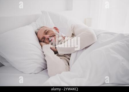 Seniorenmann schläft friedlich im Bett mit weißer Federdecke bei Tageslicht in einem gemütlichen Schlafzimmer Stockfoto