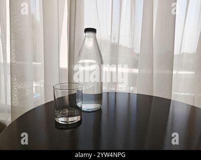 Eine Flasche Wasser steht neben einem Glas auf einem dunklen Holztisch in einem sanft beleuchteten Raum mit hellen Vorhängen. Stockfoto