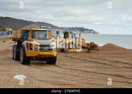 Ein Volvo A40E Kipper, der Kieselsteine entlang des Hythe Beach in der Nähe von Folkestone, Großbritannien, transportiert. Ein caterpillar-Bagger hinter dem Lkw bewegt die Schindel in Position. Der Schindel wird jedes Jahr recycelt und bewegt den Schindel entlang der Küste von Ost nach West, um Überschwemmungen und Küstenerosionen zwischen Folkestone und Hythe zu reduzieren. Stockfoto