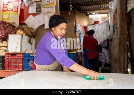 Lurin, Peru — 6. September 2024: Junge Frau in einem lila Hemd und Küchenschürze reinigt einen weißen Tisch in der bescheidenen Gemeinschaftsküche Stockfoto