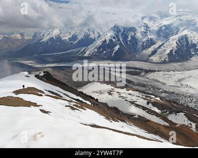 Der Wanderer steht auf einem schneebedeckten Berggipfel mit Blick auf das weite Gletschertal, umgeben von hohen Gipfeln und Wolken. Stockfoto