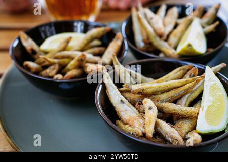 Knusprig gebratener kleiner Fisch, serviert mit Zitronenscheiben in dunklen Schüsseln auf einem Holztisch, begleitet von einem Glas Bier im Hintergrund. Ein zwangloser Appetit Stockfoto