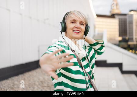 Glückliche Reife Frau in grün und weiß gestreiftem Pullover, der Musik mit Kopfhörern genießt und in der Stadt tanzt, lächelnd und in die Kamera schaut Stockfoto