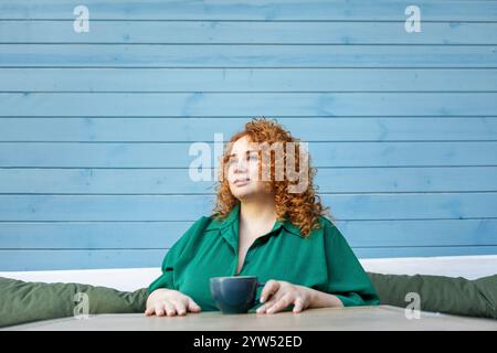 Schöne Frau in Übergröße mit rotem Haar, die eine Tasse Kaffee genießt. Kurviges weibliches Model sitzt am Cafe-Tisch in grünem Kleid gegen helles buntes blaues W Stockfoto