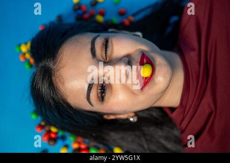 Verspielte junge Frau mit rotem Lippenstift, die ein gelbes Bonbon zwischen den Lippen hält und auf einem blauen Hintergrund mit bunten Bonbons zwinkert Stockfoto