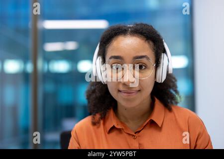 Eine professionelle Frau mit Kopfhörern und Brillen im Büro, die konzentriert und selbstbewusst wirkt. Sie trägt ein lässiges oranges Hemd, das eine moderne Arbeitsumgebung hervorhebt. Stockfoto