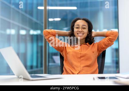 Eine selbstbewusste Geschäftsfrau in einem orangefarbenen Hemd entspannt sich an ihrem Schreibtisch in einem modernen Büro und lächelt in die Kamera. Die Szene vermittelt Professionalität, Komfort und ein positives Arbeitsumfeld. Stockfoto