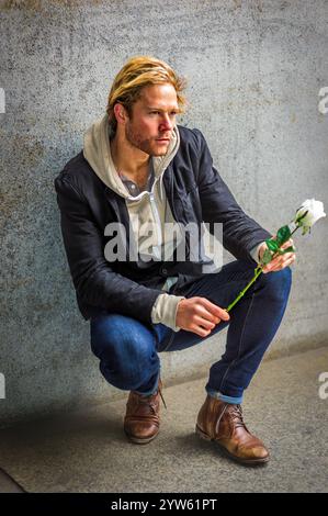 Ein Mann mit blonden Haaren hockt sich an einer Betonwand, hält eine weiße Rose und sieht besinnlich aus. Er trägt eine schwarze Jacke, eine Kapuze Stockfoto