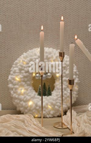 Weihnachtliche Einrichtung, weicher Plüschkranz mit Lichtern, beleuchtete Kerzen in goldenem Halter, beigefarbenes Tuch auf Holztisch Stockfoto