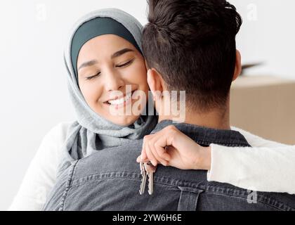 Emotionale junge Dame in Hijab mit Schlüssel umarmt ihren Mann Stockfoto
