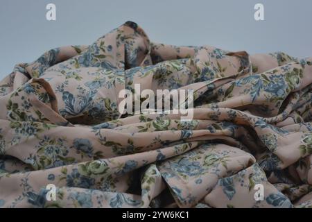 Ungewöhnlicher, glänzender Sommerstoff. Beige, dünne synthetische Seide mit blauen Blüten und grünen Blättern auf weißem Kunststoffhintergrund. Stockfoto