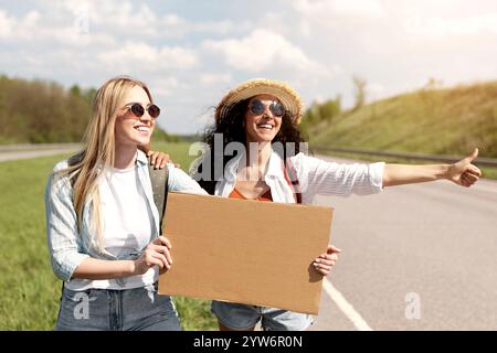 Zwei Freundinnen, die eine Freifahrt anhängen, ein leeres Kartonschild mit Mockup halten, das Auto auf der Straße anhalten, freien Platz Stockfoto