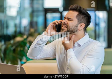 Ein kranker Geschäftsmann, der über das Smartphone spricht, Halsschmerzen hat, Husten hat und mit einem Laptop arbeitet. Gestresster müder Mann, der an Erkältung oder Grippe litt und in der Lobby des Büros arbeitet. Trauriger Arbeiter hat Virus, Krankheit. Stockfoto