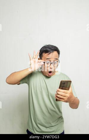 Porträt eines überraschten asiatischen Mannes mit einer Brille, der ansieht, was er auf seinem Smartphone vor weißem Hintergrund der Studiowand sieht, Mann in hellgrünem T-shir Stockfoto