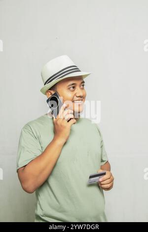 Erwachsener asiatischer Mann mit Strohhut und hellgrünem Hemd, entgegennehmendem Anruf auf seinem Handy, Seitenansicht mit Karte und lächelnd glücklich. Reisen Stockfoto