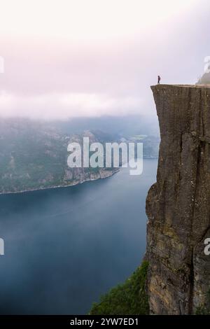 Ein einsames Individuum steht am Rande von Preikestolen, einer Klippe in Norwegen mit Blick auf einen Fjord. Der Himmel ist nebelig und dramatisch. Preikestolen, Norwegen, Konz Stockfoto